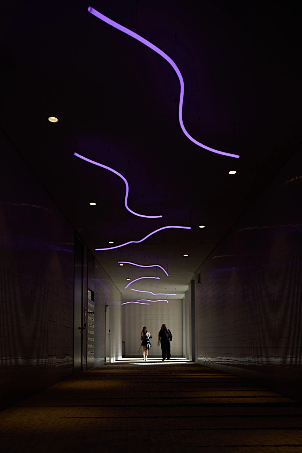Dark corridor, with neon shapes and people passing through it.